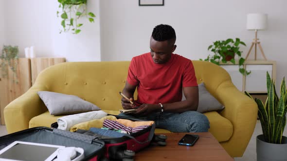 Young Adult Man Packing Clothes to the Suitcase and Writing Travel Checklist