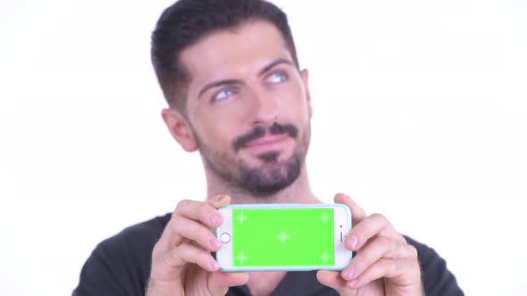 Face of Happy Young Handsome Bearded Man Thinking While Showing Phone
