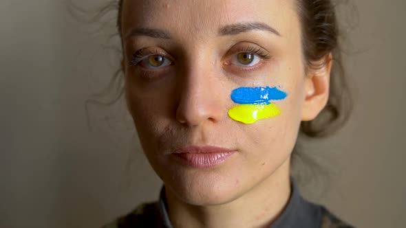 Indoor Portrait of Young Girl with Blue and Yellow Ukrainian Flag on Her Cheek Wearing Military