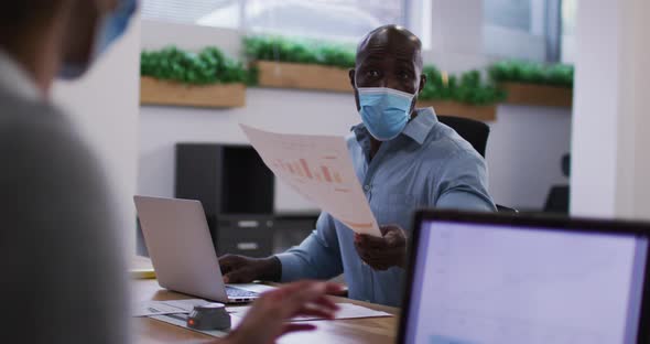Diverse male and female business colleagues in face masks using laptops passing document in office