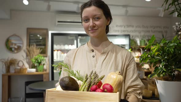 Smiling Woman Holds Box of Vegetables and Looks at Camera