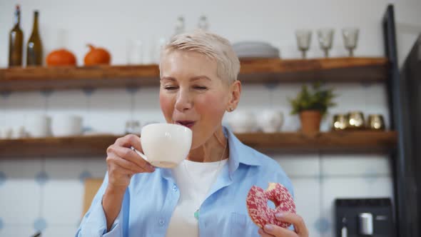 Smiling Senior Woman Drinking Coffee and Eating Delicious Glazed Donut at Home