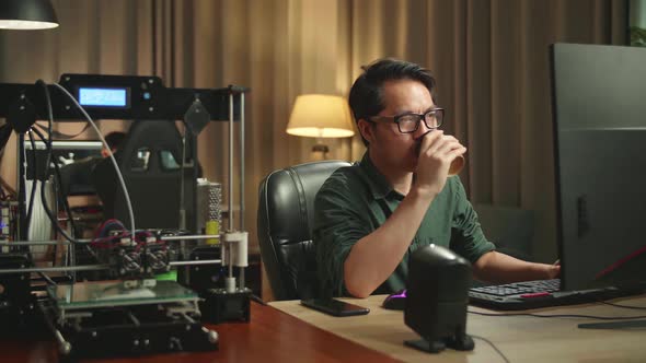 Asian Man Drinking Coffee While Works On Personal Computer And 3D Printer In Home Office