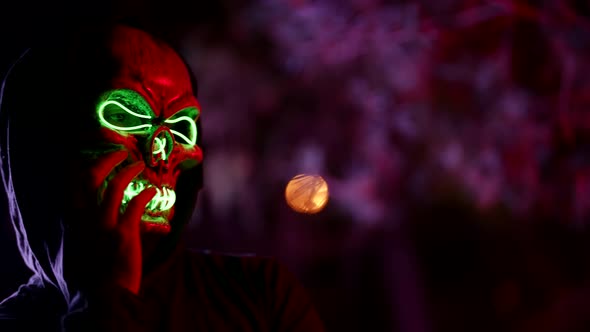 Portrait of a Person in a Terrible Glowing Death Mask and a Hood on a Blurry Reddish Background with