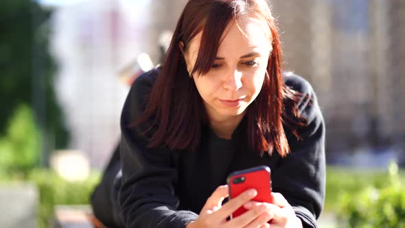 Young Woman Lying on Her Stomach Outside on a Bench with Her Legs Up, and Browsing Her Cell Phone. A