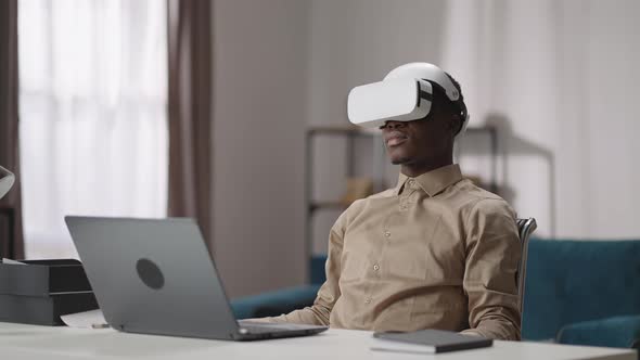 Young Black Man is Using Headmounted Display for Playing Video Games Sitting at Table with Laptop