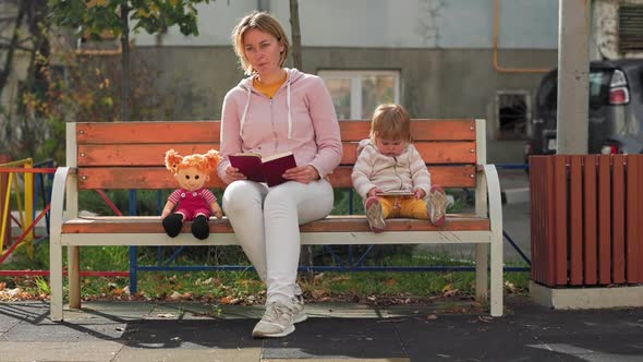 Young caucasian mother is reading a book sitting on a bench.