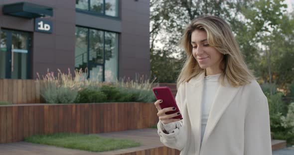 Attractive Young Woman Using Smartphone While Smiling and Walking at City Street. Stylish Female