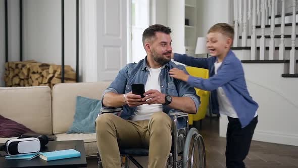 Man in Wheelchair Using His Smartphone and Hugging His Cheerful Small Son which Running to Him