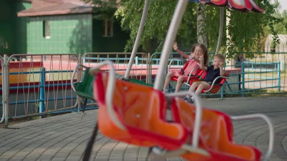 Happy Little Children Wave Hands Riding Carousel in Park