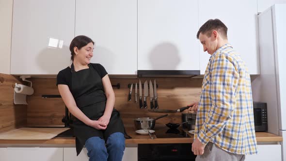 Man Turns a Pancake in a Frying Pan and a Woman Sits on Table Next