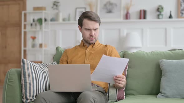 Young Man with Laptop Reacting to Loss on Documents, Sofa