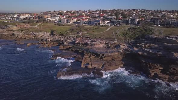 Flying sideways aerial shot of ocean waves crashing at seashore and rockpool near waterfront houses