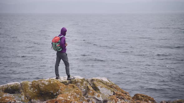 Traveler Young Man with Colorful Backpack is Raising His Arms in Front of Stormy and Windy Sea