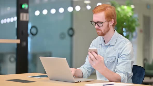 Young Redhead Man Using Smartphone and Laptop in Office