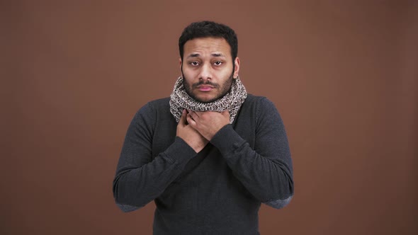 Sick Indian Man Having Sore Throat Wearing Scarf and Touching His Neck Coughing Over Brown Studio