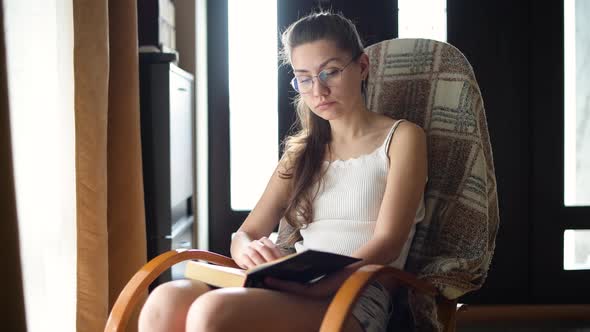 Young girl in glasses reads a book in natural light on a rocking chair