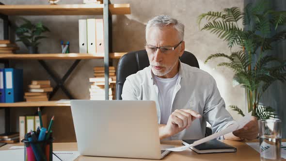 Middleaged Man Saying Hi to Someone Using Online Video Call on Laptop Showing Diagram on Sheet of