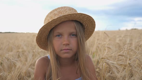 Portrait of Little Girl in Straw Hat Looking Into Camera Against the Background of Wheat Field at