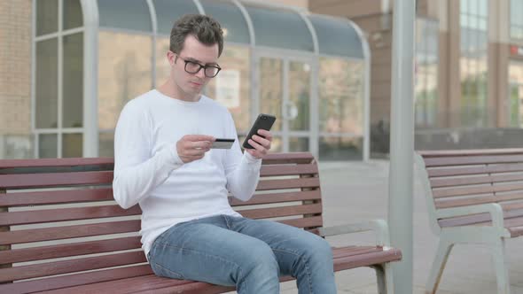 Excited Young Man Shopping Online Via Smartphone While Sitting Outdoor on Bench