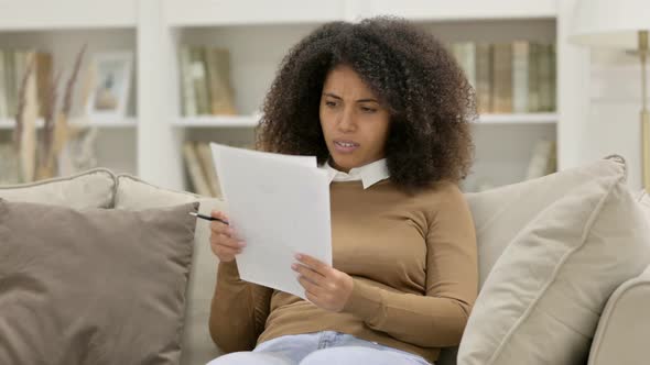 Young African Woman with Documents Reacting To Loss on Sofa