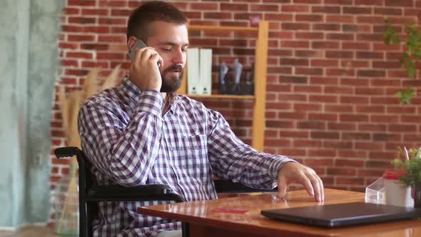 Person with Disabilities is Sitting on Wheelchair at Table with Laptop and Talking on Phone