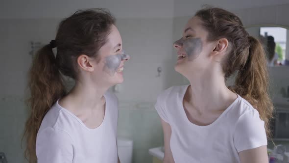 Two Slim Beautiful Identical Women with Cosmetic Facial Mask Laughing Looking at Each Other Turning