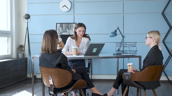 Three business girls are sitting on chairs at a table drinking coffee and discussing new projects