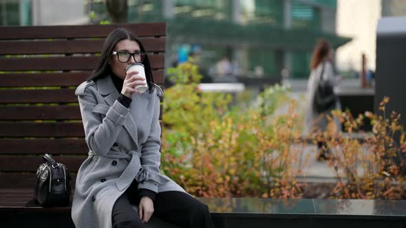 Adult Woman in Grey Coat is Drinking Coffee From Carton Cup in Downtown