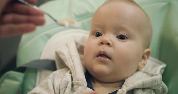 Cute Toddler in Kid Chair During Feeding with Spoon at Camera