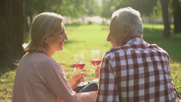 Merry Senior Couple Sitting on Grass in Park and Drinking Wine, Celebrating Date