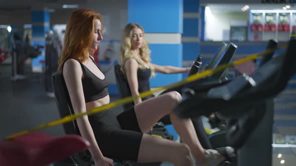 Side View Portrait of Slim Redhead Caucasian Woman Riding Exercise Bike with Blond Friend Training
