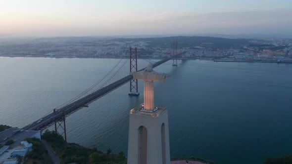 Aerial Evening Back View of Big Jesus Statue on Pedestal and Long Bridge Over River