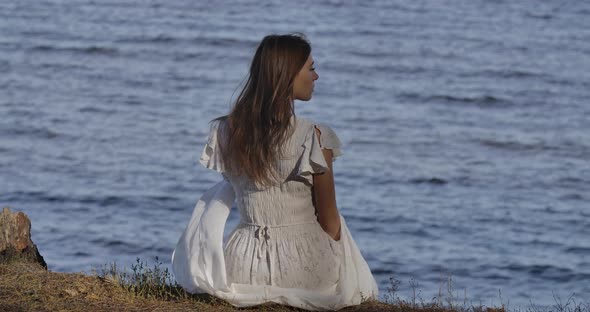 Back View of an Elegant Caucasian Woman in Light White Dress Sitting on the Beach and Thinking