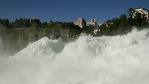 Zoom in shot of splashing rhine water fall with swiss flag on mountain and riding train in backgroun