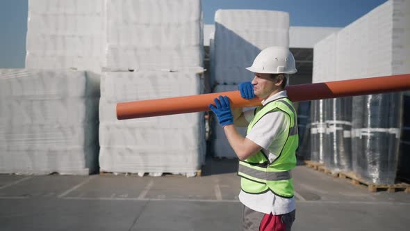 Side View Strong Caucasian Man in Hard Hat Walking with Orange Pipe on Shoulder in Slow Motion