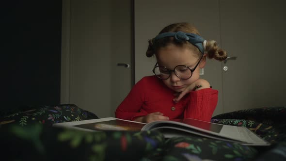 Little Girl in Glasses Lying on the Bed and Reading Book