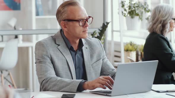 Mid-Aged Businessman Typing on Laptop at Desk in Open Space Office