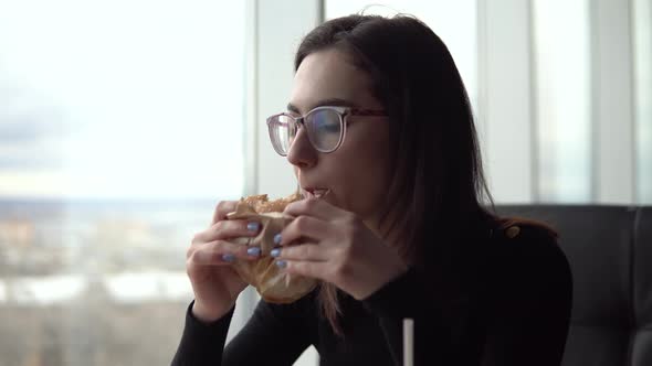 A Young Woman Is Eating a Burger. A Girl Sits in a Cafe By the Panoramic Window and Eats Fast Food.