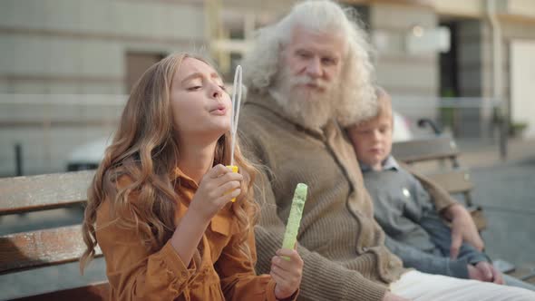 Camera Approaches To Happy Little Girl Blowing Soap Bubbles with Blurred Grandfather and Brother