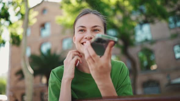 Laughing blonde woman wearing green t-shirt recording voice message by phone on the bench