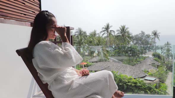 Asian woman sitting on a balcony, wearing white dressing gown and drinking coffee while looking at p
