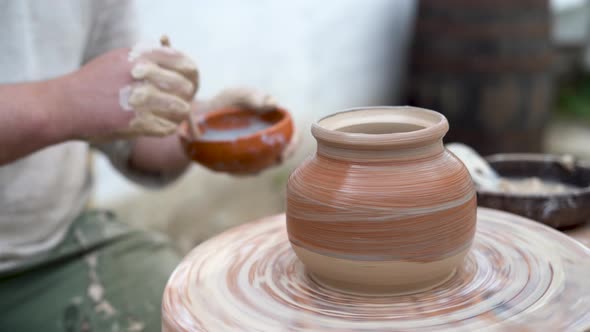 The Master Potter Paints a Clay Jug with White Paint on an Old Potter's Wheel Twists His Foot