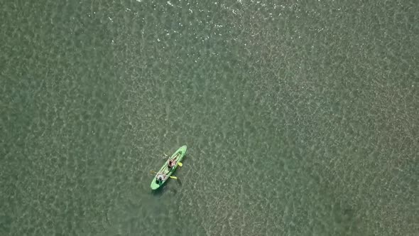 two people in sit-on-top kayak paddle in shallow green water, aerial
