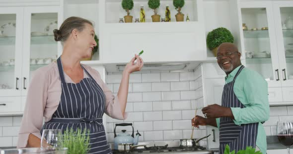 Smiling senior diverse couple wearing blue aprons and cooking in kitchen