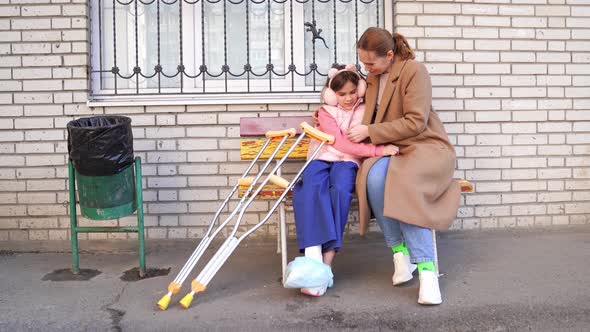 Mom with Daughter with Crutches Sits on Bench in Yard of Apartment Building