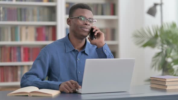 Young African Man Talking on Phone While Using Laptop in Office