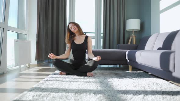 Young Caucasian Woman in Black Jumpsuit Sitting in Lotus Position and Meditating Yoga at Home