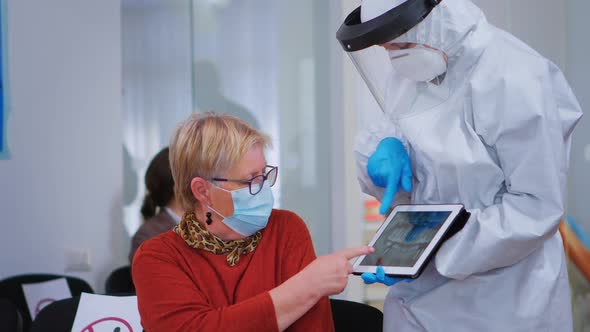 Woman Sitting on Chair Listening Doctor with Coverall Looking on Tablet