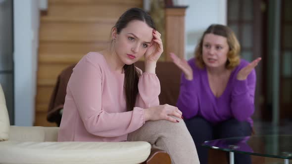 Overwhelmed Caucasian Young Woman Sitting at Home As Angry Mother Shouting Gesturing at Background